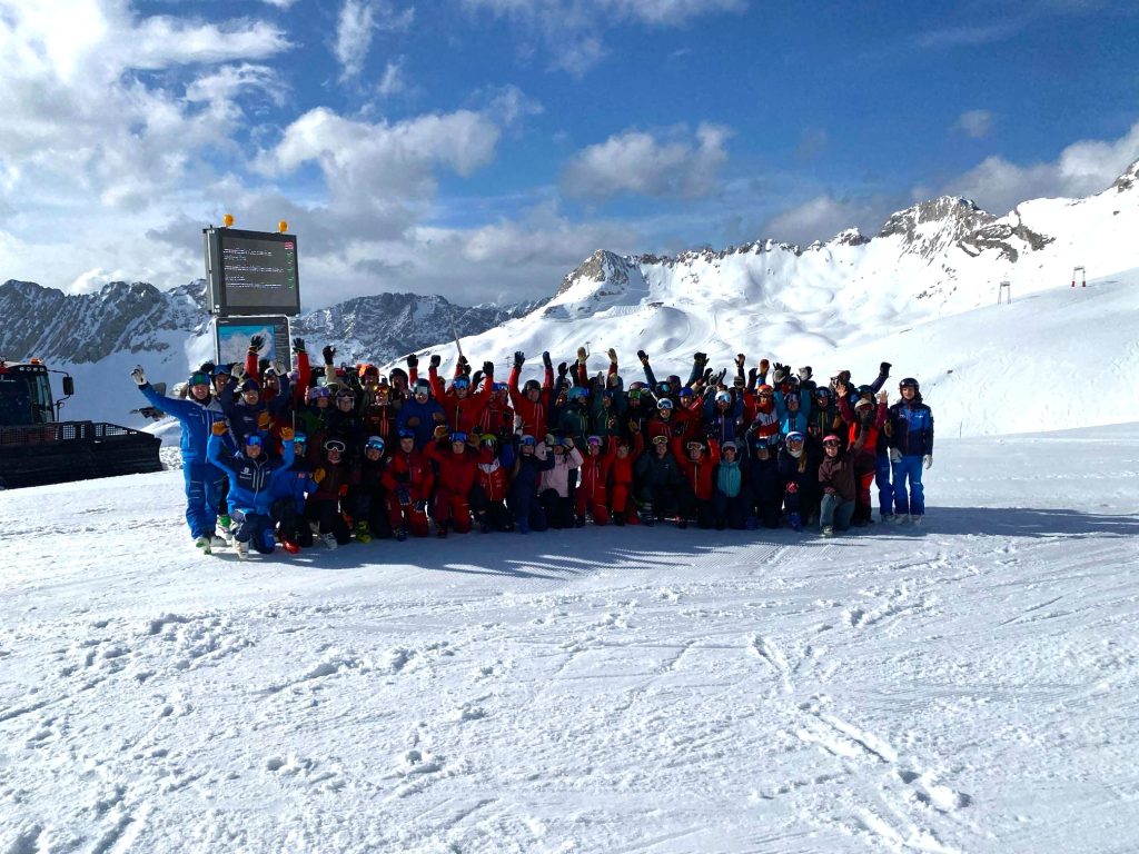 Eine große Gruppe von Skifahrern in bunter Skibekleidung posiert gemeinsam lachend auf einer sonnigen Skipiste vor einer verschneiten Alpenkulisse. Alle heben jubelnd die Arme, im Hintergrund sind schneebedeckte Berge zu sehen.