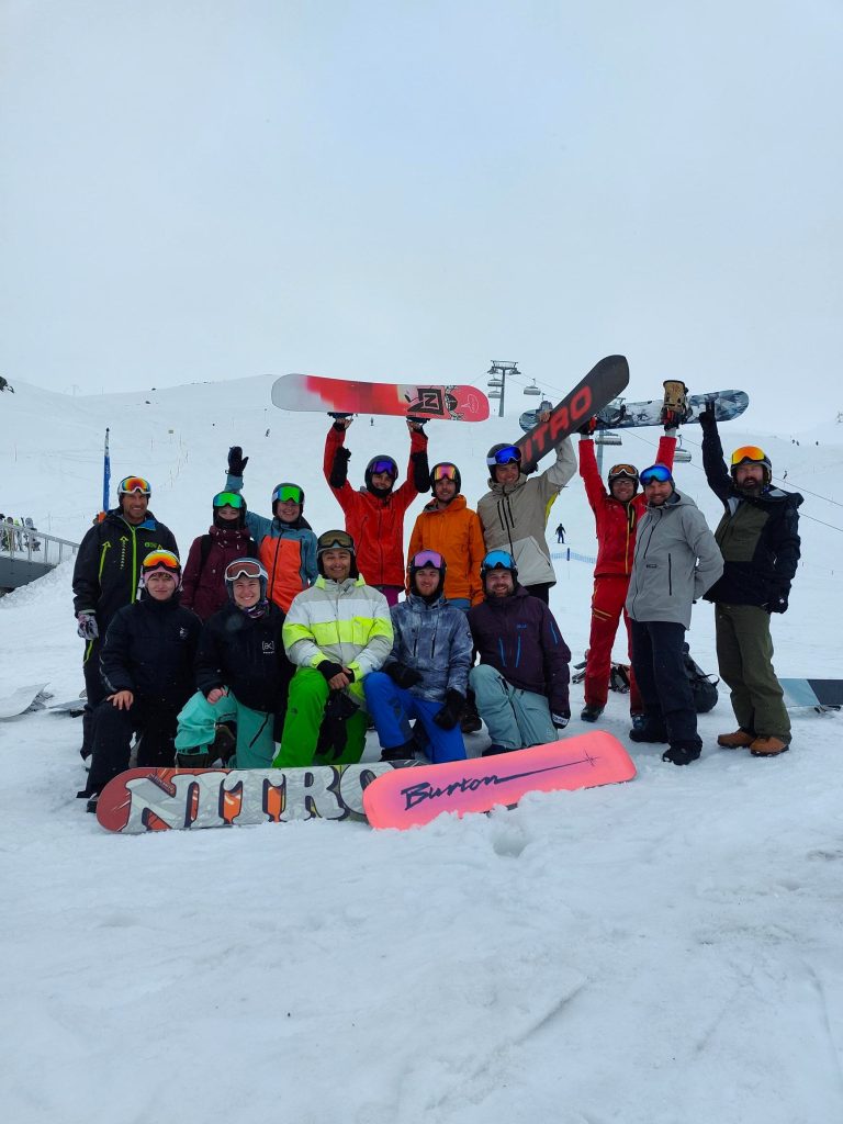 Gruppe fröhlicher Snowboarder vor verschneiter Alpenlandschaft.