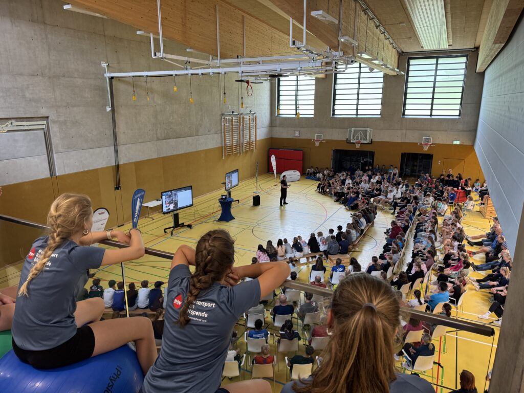Bild von einer Sporthalle innendrinnen mit Personen auf Stuhlreihen