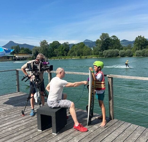 Interview Team mit Kamera und Microfon interviewt Wakeboarder vor Wasserskianlage