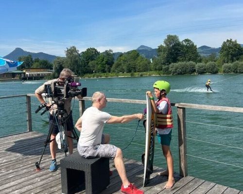 Interview Team mit Kamera und Microfon interviewt Wakeboarder vor Wasserskianlage