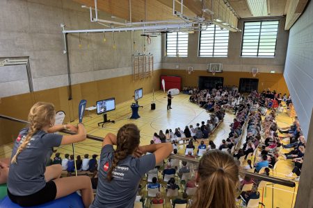 Bild von einer Sporthalle innendrinnen mit Personen auf Stuhlreihen