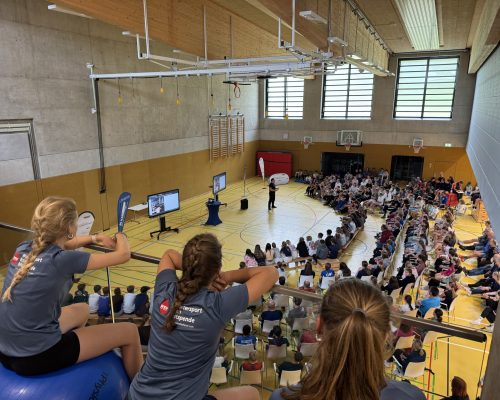 Bild von einer Sporthalle innendrinnen mit Personen auf Stuhlreihen